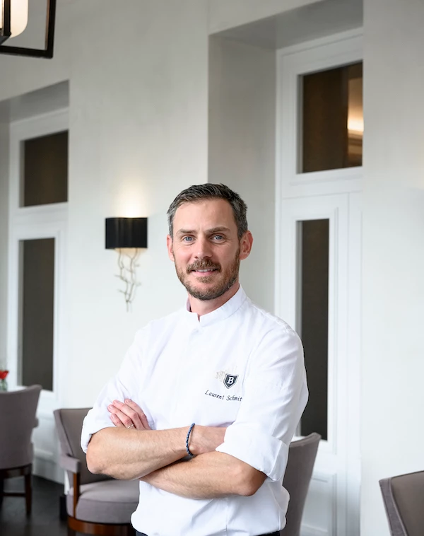 Professional chef in a crisp white uniform stands confidently in an elegant restaurant, offering an inviting and competent air perfect for culinary connoisseurs.