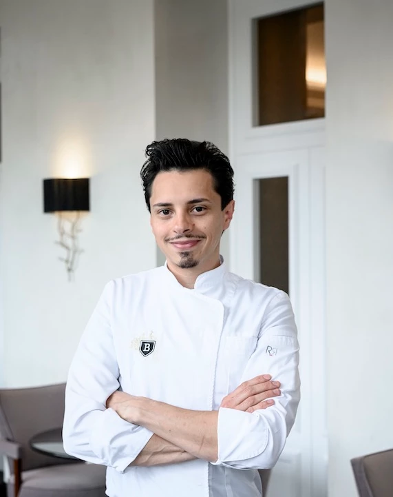 Confident chef in a white uniform stands in a minimalist dining area, exuding professionalism in the culinary industry.