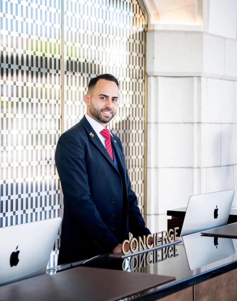 Professional concierge in a blue suit by a desk with computers, set against a tiled backdrop, projecting efficiency and hospitality in the service industry.