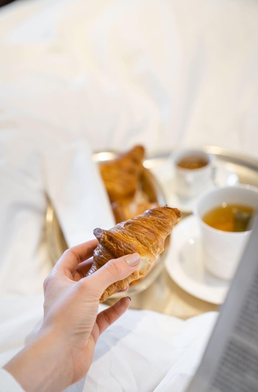 Woman holding a flaky, golden croissant over a soft white bedsheet, with tea in the background; evokes a cozy, relaxed morning.